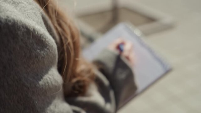 Photographer notes environments using pen during daytime reporting session. Female correspondent records spontaneous observations with blue pen on spiral notebook in bright sunlight