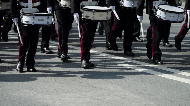Uniformed snare drummers in a marching band stride in tight formation along a city street, white gloves gripping drumsticks as rhythmic footsteps echo during a parade performance