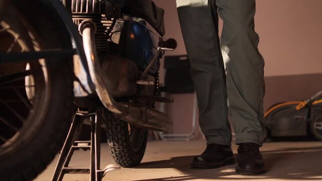 Man wearing work pants and sturdy boots placing his foot on the kickstart lever of a classic motorcycle, preparing to start the engine for a ride or repair in a home garage