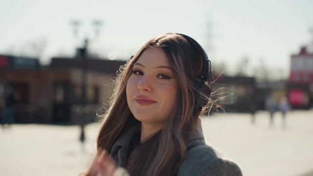 Casual woman with headphones enjoying city scene. Cheerful Asian woman in grey coat listening to music outside. Influential woman with playful glance and warm smile in urban outdoor setting