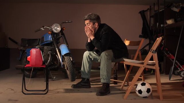Man covers his face in frustration while sitting in a garage watching a soccer match on an old tv, surrounded by a motorcycle and a football, feeling isolated and disappointed