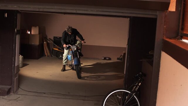 Man sitting on a vintage blue motorcycle inside a cluttered garage with the door open, preparing for a ride and ready to start a new journey or adventure