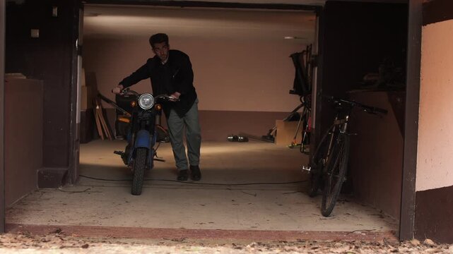 Man pushing blue vintage motorcycle out of dim residential garage, moving and maintaining a classic bike in a cluttered workshop like storage space, casual weekend project