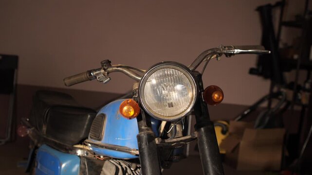Blue vintage motorcycle, featuring a prominent round headlight and handlebars, sitting in a garage, symbolizing a hobby project, restoration, or forgotten classic transport