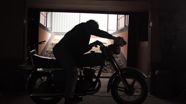 Man in silhouette working on a classic old timer motorcycle in a dark garage, preparing the machine for a ride while performing maintenance in a home workshop
