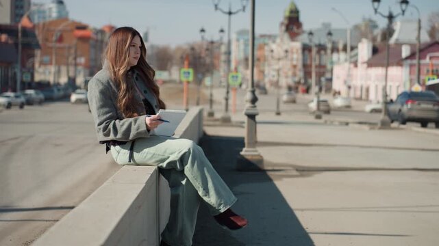 Pretty white woman writing travel journal on bench sunlit city plaza with historic architecture and lamp posts, careful handwriting on notebook, reflective mood, solitude and observation, cinematic