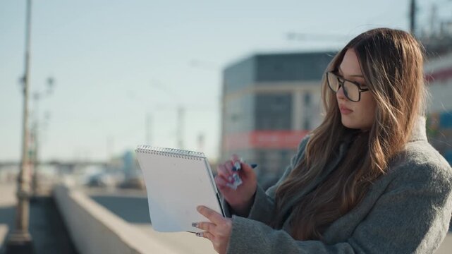 Caucasian woman creating detailed urban sketch. Woman in coat sketching at sunny city plaza with glasses. Focused female artist drawing on park bench amidst bright winter sunlight in busy city area