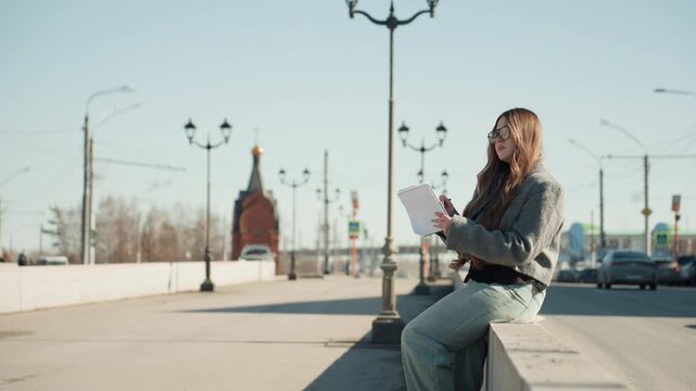 Confident woman examines sketches outdoors. Stylish woman reviewing artwork in city environment. Fashionable woman in gray coat analyzing creative drawings against bright city background