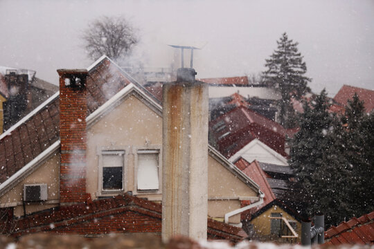 Smoke billows from a modern brick chimney on snow covered tiled roof. Exterior view of house rooftop with heating pipe emitting dark fumes, urban background. Belgrade Zemun Serbia Snowfall Blizzard