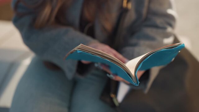 Pretty turquoise notebook in hands caucasian woman student sits on bench in gray coat, flipping pages and jotting notes with pen closeup on textured leather cover, stitched binding and paper edges,
