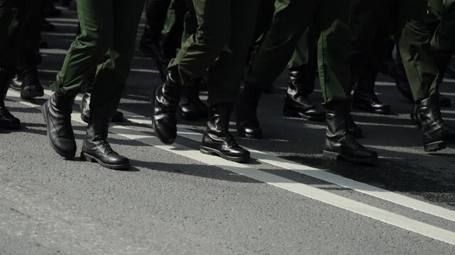 Close-up of uniformed personnel marching in tight formation, black combat boots and green trousers on asphalt beside white lane lines, showing discipline and synchronized movement.