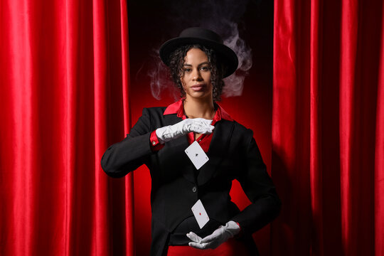Young African-American magician showing magic trick with flying cards near red curtains on dark background