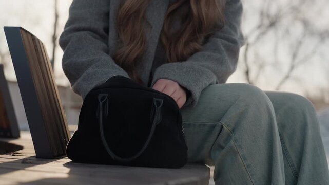 Pretty seated woman adjusts black purse on bench during golden winter light, commuter pauses with hands on zipper, long hair and wool coat frame thoughtful pose, knit trousers and muted palette create