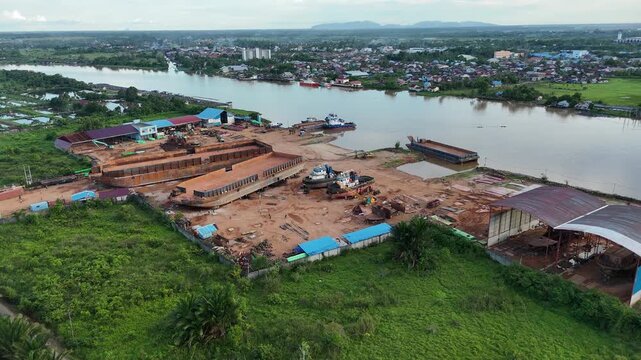 An aerial view of a shipyard in operation, featuring large rusting ships and barges under construction on dry land along the banks of the Kapuas River in West Borneo, Indonesia.