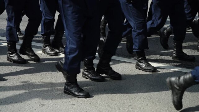 Close-up of uniformed personnel marching in synchronized formation along a paved street, black boots and blue trousers moving in rhythmic unison across double white lane markings.