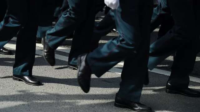 Close-up of uniformed marchers in synchronized formation, teal trousers with red piping and polished black shoes stepping in unison along sunlit asphalt during a disciplined parade
