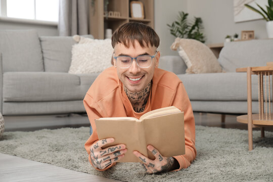 Young tattooed man in eyeglasses reading book on floor at home