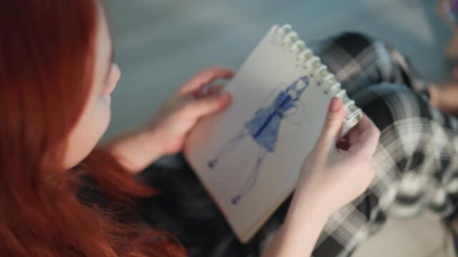 Redhead woman holding blue dress sketch on lap, plaid trousers visible, delicate pen strokes define silhouette, reflective posture suggests stylist study or model casting, soft natural light