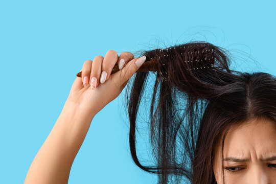 Beautiful young stressed Asian woman trying to brush tangled hair on blue background, closeup