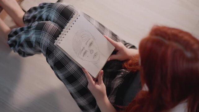 Redhead caucasian woman studying face chart overhead, sketchbook portrait on lap, careful finger tracing of features, graphite linework, plaid trousers on wooden floor, focused creative practice