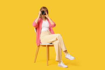 Beautiful young woman sitting on chair with modern camera on yellow background © Pixel-Shot
