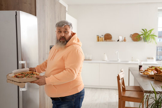 Shocked overweight mature man with pizza near fridge in kitchen. Overeating concept