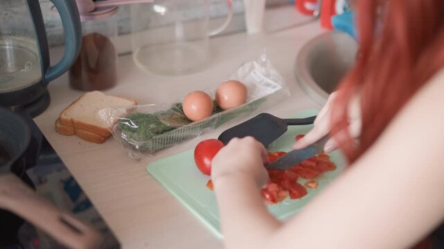 Redhead white woman chopping tomato on mint cutting board, eggs and loaf of bread beside herb packet, soft morning light, calm hands, mindful breakfast preparation, cozy domestic ritual with subtle