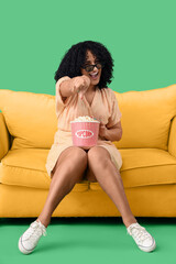 Obraz premium Beautiful woman in 3D glasses with popcorn on sofa pointing at viewer against green background