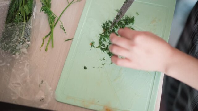 Hands chopping fresh herbs on cutting board, closeup showing herbalist slicing parsley and cilantro with small chef knife, scattered stems and plastic wrap on counter, focused mincing motion, warm