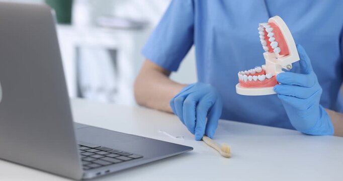 Dentist in medical gloves showing how to clean teeth with model of jaw during online consultation by laptop at white table in clinic, closeup