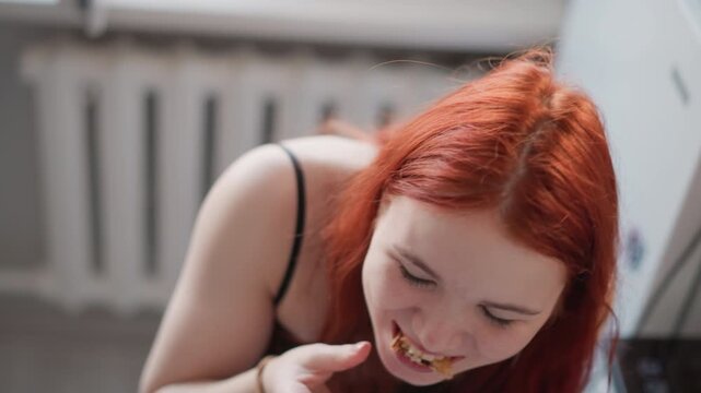 Young woman eating sandwich with gusto, energetic messy devour shows expressive face, crumbs and filling falling, casual apartment setting with radiator background, high energy bite conveying hunger,