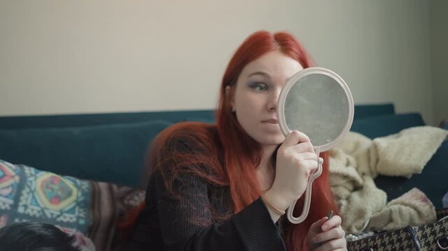 Redhead caucasian woman on couch holding small mirror, applying makeup and inspecting earrings with cozy blanket and patterned pillows frames portray makeup artist practicing technique, private seer