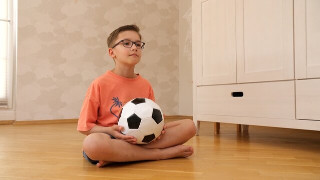 Little boy wearing eyeglasses and an orange t shirt holding a soccer ball, sitting cross legged on a wood floor at home, patiently waiting for playtime or activity in childhood