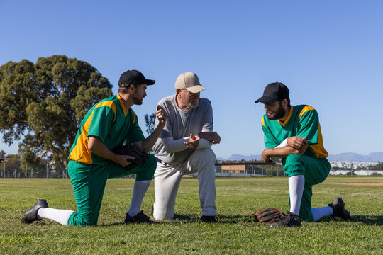 Senior coach coaching two male players kneeling on local ballfield in green uniforms with gloves