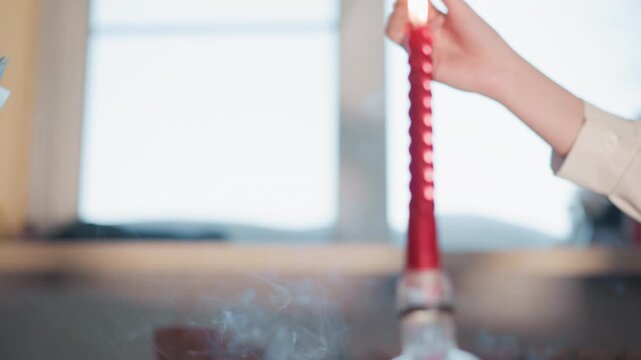 Hands exchanging talismans beside red candle, incense smoke curling upward, closeup of reader and client hands passing metallic charms and small card, lit candle on wooden table, soft window light,