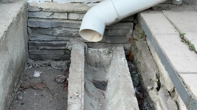 White pipe over concrete drain channel closeup view of elbow spout above empty concrete gutter beside brick wall, dry basin