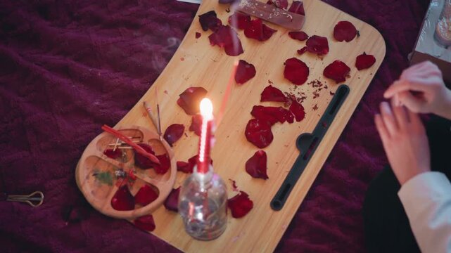 Candlelit fortunetelling setup with rose petals, mystic reader arranging petals on wooden board beside lit candle on velvet cloth, small bowl with herbs and crystal, hands performing focused reading