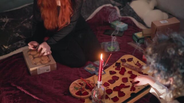 Candlelit floor reading with heart board, redhead fortune teller arranges heart tokens on wooden board beside tall red candle, client watches from edge of velvet blanket, incense smoke curls, intimate