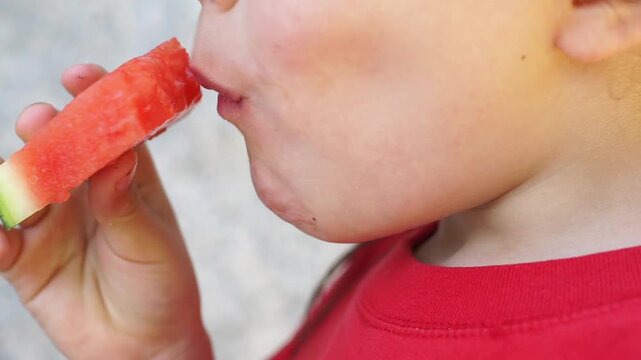 A close-up shot of a young child happily biting and chewing a fresh, red slice of watermelon on a bright summer day.