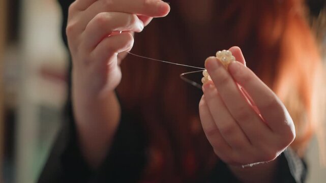 Closeup caucasian hands untangling charm, soft light, red hair silhouette, delicate beads on thread, focused fortune teller preparing talisman, intimate parlor atmosphere, mysticcore mood, prediction