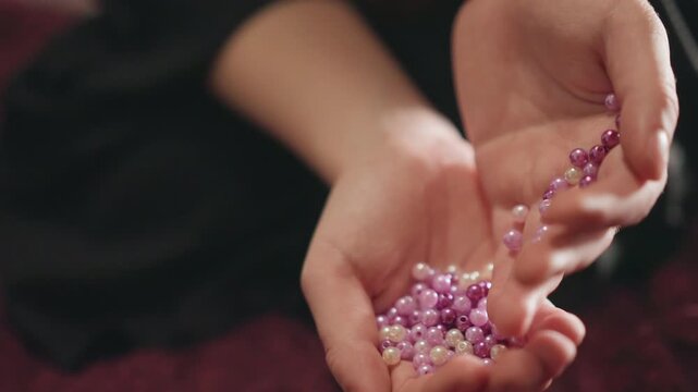 Closeup caucasian hands pouring purple pearls into palm, workshop table, careful counting motion, jewelry artisan at work, tactile texture, cozy slowliving aesthetic, focused craft mood, subtle