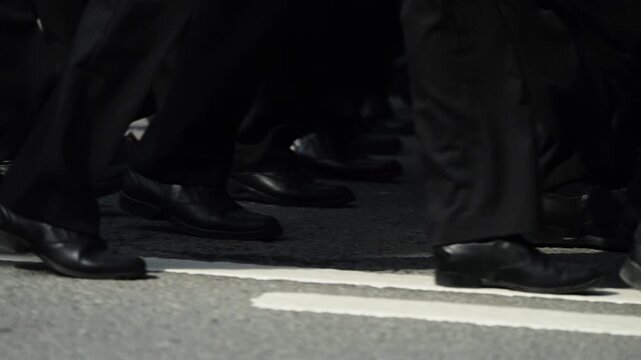 Close-up of synchronized marchers in black trousers and dress shoes stepping in unison across painted street lines, highlighting movement and rhythm on an urban road