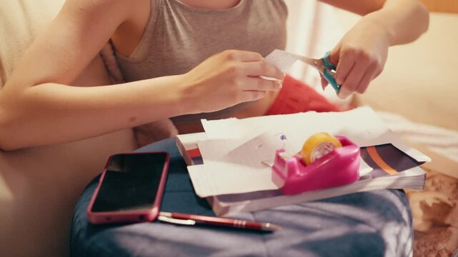 Close up of a focused student hand using adhesive tape and scissors on a small piece of paper, finishing a secret crib sheet and notes for a test while sitting on a sofa in a living room.