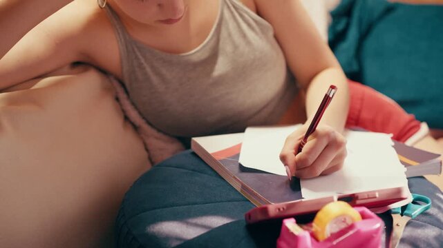 Close up of a concentrated teenage girl copying small text from her phone to create a tiny cheat sheet, preparing for an academic exam while sitting on a comfortable sofa in a living room.
