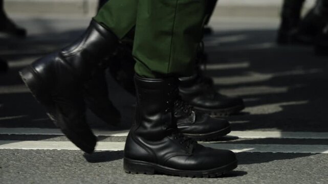 Close-up view of uniformed personnel marching in synchronized formation, black combat boots striking the asphalt in rhythmic steps, green trousers moving in unison along a roadway.