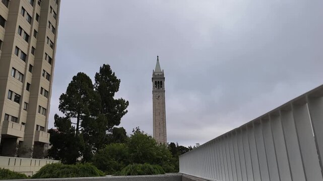 Overcast sky with clouds moving over the Sather Tower, also known as the Campanile, at the University of California, Berkeley, an iconic landmark surrounded by trees and modern buildings