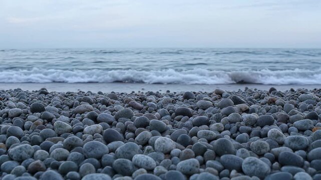 Gentle waves lap against a beach of smooth grey pebbles with a soft blue sky and ocean horizon.