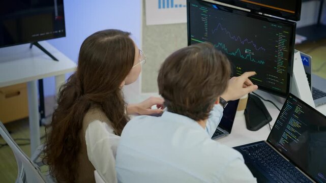 Female data scientist and male software engineer reviewing live machine learning training dashboard and GPU monitoring on screen. Artificial intelligence research, data analysis, technology workspace.