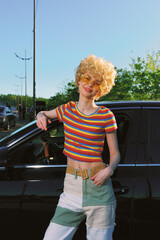 Young woman with curly blonde hair leans on a dark car wearing a bright striped crop top and...
