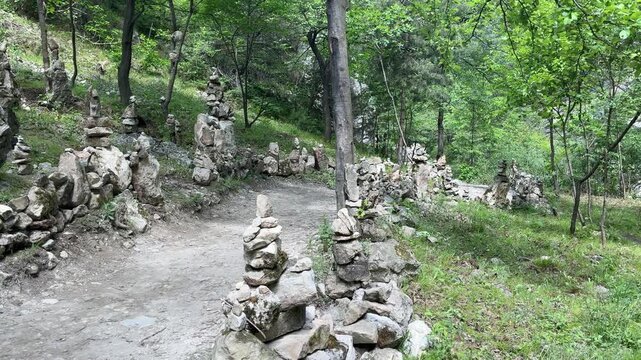 Numerous rock cairns line a dirt path through a lush green forest on a sunny day in Gunibsky District, Republic of Dagestan, Russia